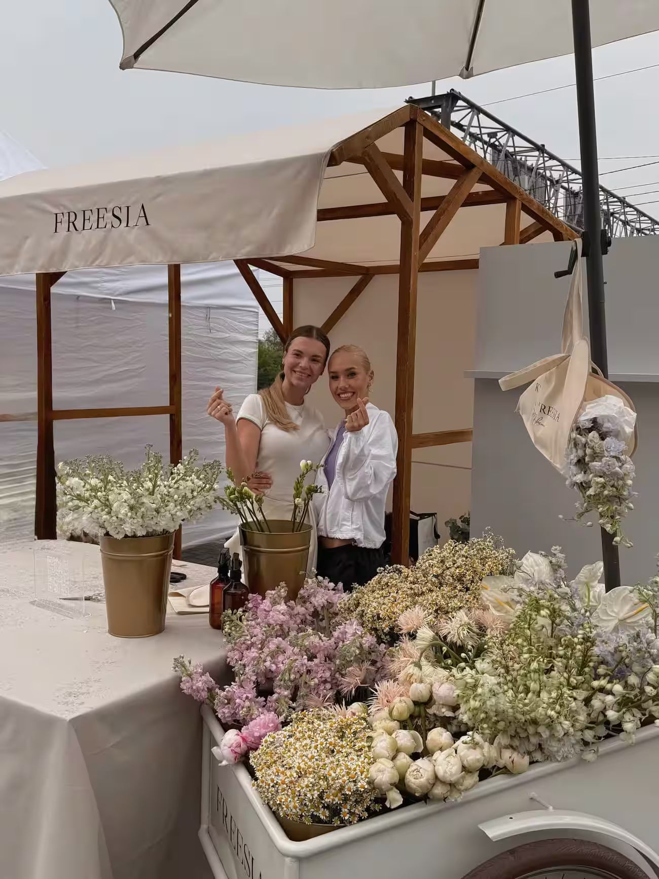 Flower market stall with fresh bouquets, two women smiling, outdoor setting, soft pastel flowers